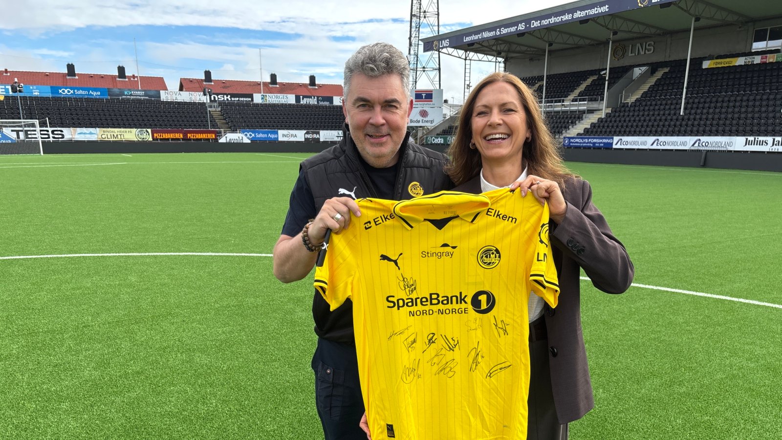 Frode Thomassen og Nina Hovland holder opp en Glimt-drakt ute på Aspmyra stadion. 