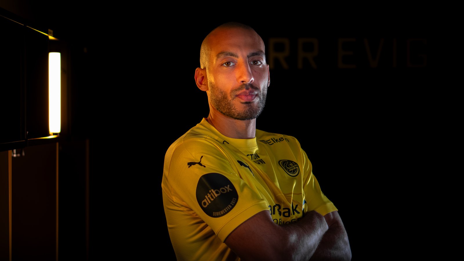 En smilende Haitam Aleesami står i et mørkt studio med Glimt-drakt.