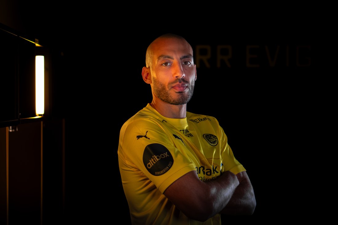 En smilende Haitam Aleesami står i et mørkt studio med Glimt-drakt. 