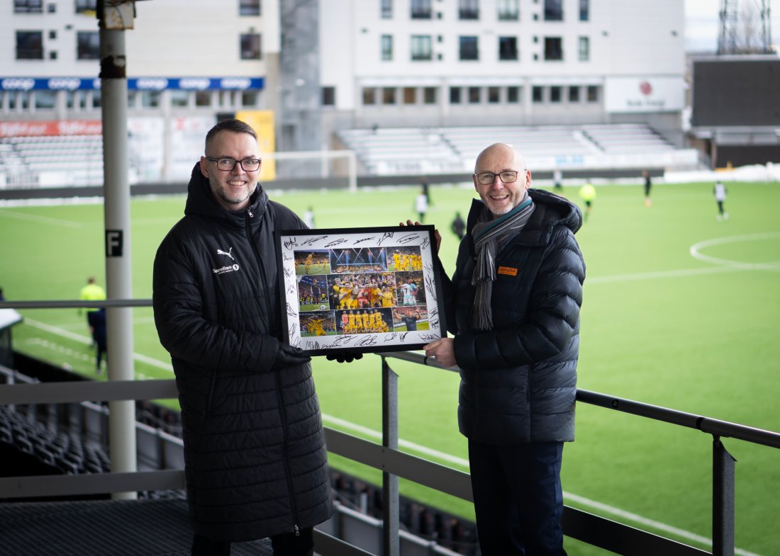 Bilde av smilende Leder marked og salg, Tom Steffensen, i Bodø/Glimt og leder for produkt og marked, Knut-Bjørnar Braathen, i Saga Mobil på Aspmyra. Mellom seg hoder de et innrammet fotocollage fra Bodø/Glimts sesong i 2025